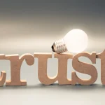 The image shows the word trust in wooden letters, with a lit light bulb resting on top of the U symbolizing the connection between trust and bright ideas or innovation.